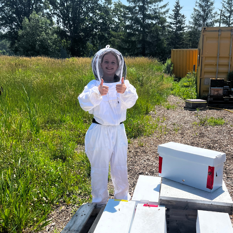 Person in a beekeeping suit giving thumbs up outdoors with bee boxes in the background