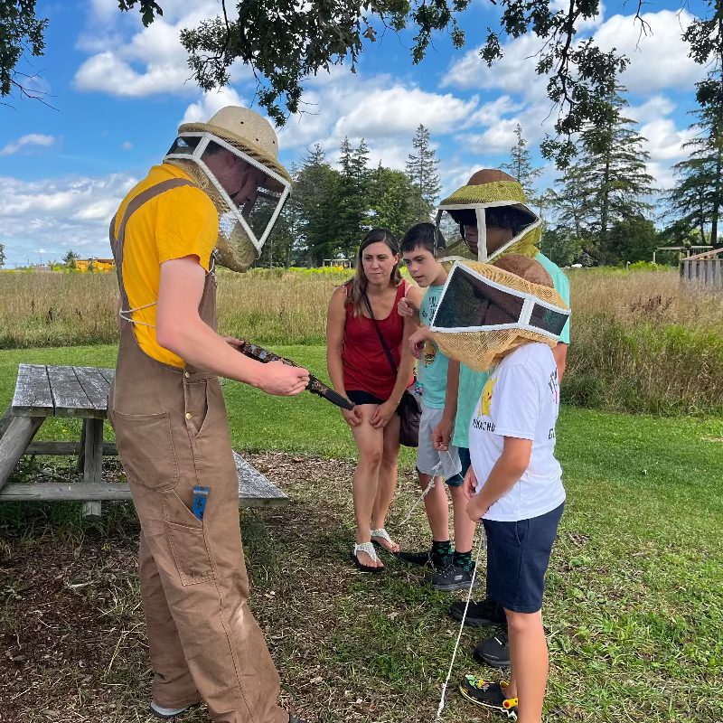 Person in beekeeping suit demonstrating to a group outdoors with picnic table and trees in background