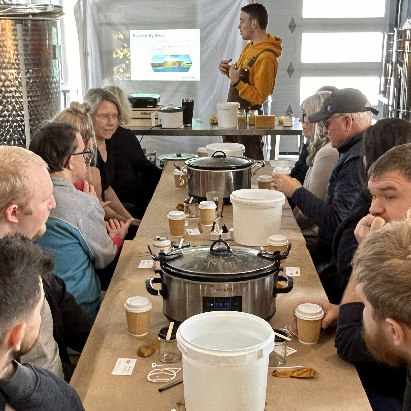 Group of people gathered around a table with cooking equipment and food items, possibly in a workshop or educational setting.