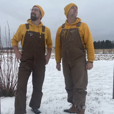 Two men wearing yellow hoodies and brown overalls standing in a snowy field.