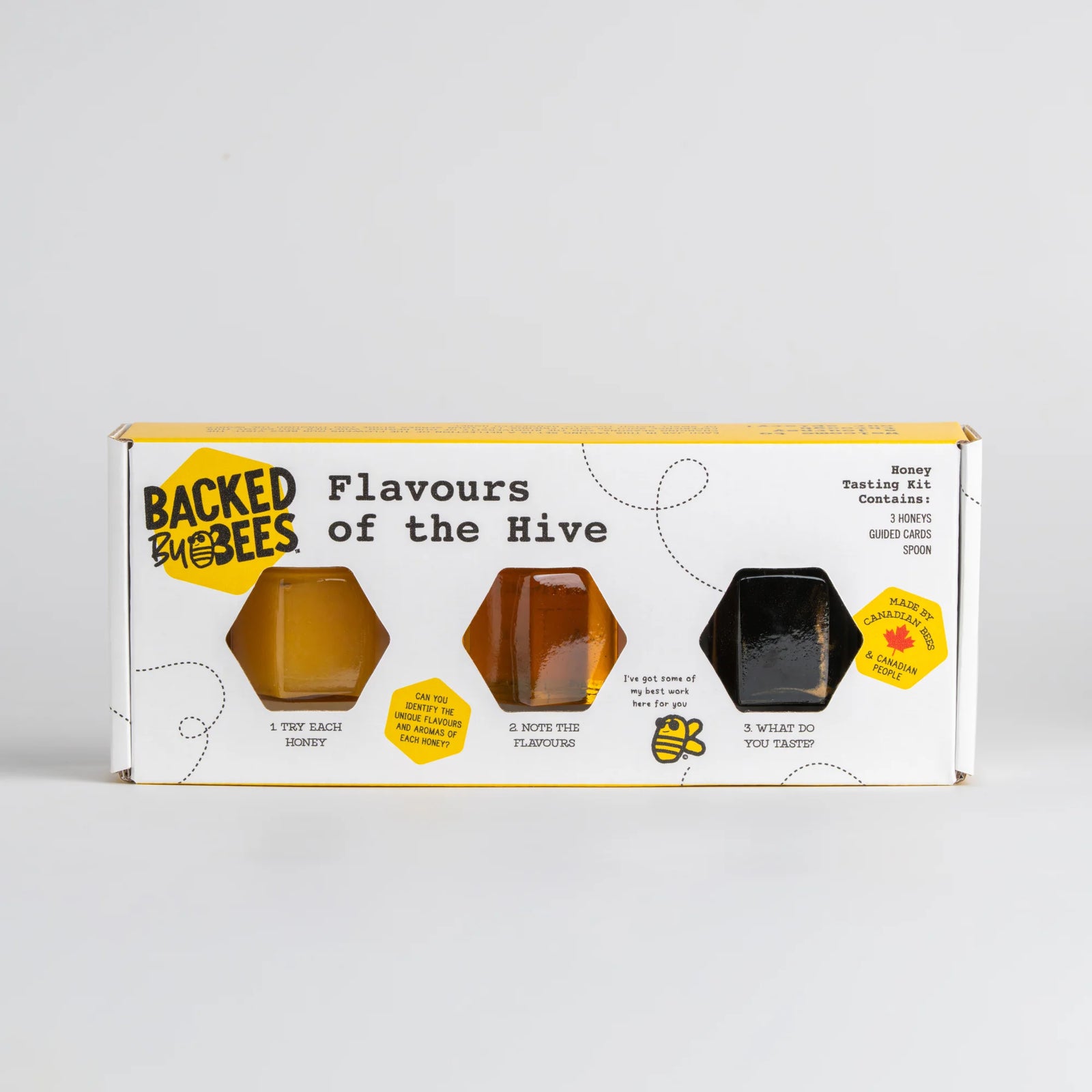 A boxed honey tasting kit from Backed by Bees, featuring three hexagonal honey samples in amber, golden, and black on a white box with yellow accents, photographed against a light gray background.