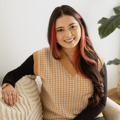 A woman with long dark hair featuring pink highlights sits on a light beige sofa wearing a tan and cream houndstooth vest over a black long-sleeve top and black pants. She is holding a textured cream pillow next to her.