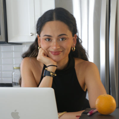 A woman with dark hair sits at a table in a kitchen setting, wearing a black sleeveless top and gold jewelry. In front of her is an open silver Apple laptop, and on the table next to her are stacked books and an orange.